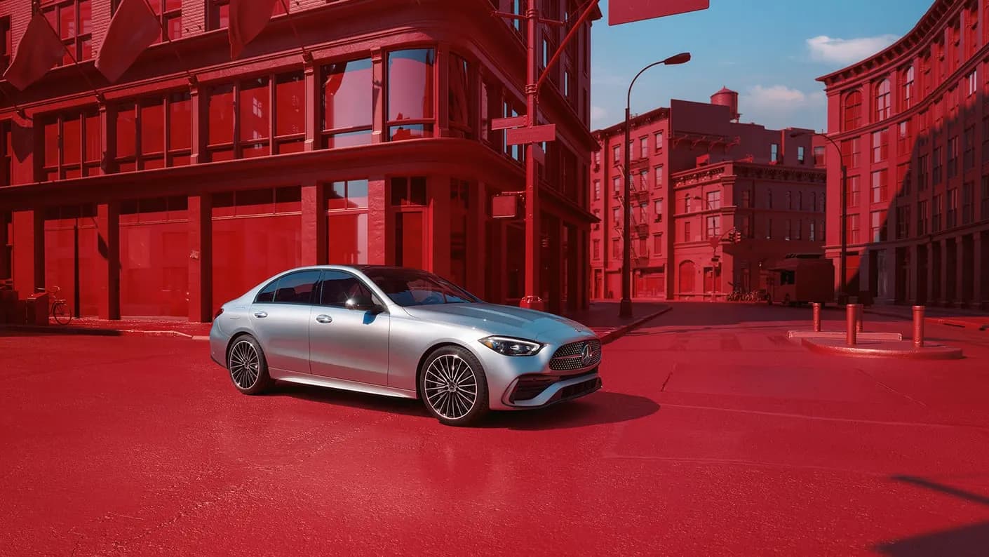 Mercedes-Benz Classe C de Kotewo en finition argent métallisé, photographiée dans un décor urbain aux tonalités rouges spectaculaires. Cette berline compacte et racée fait partie de notre flotte premium comprenant également les Mercedes Classe S, Classe E et Classe V, offrant un équilibre parfait entre élégance et agilité pour vos déplacements en région Rhône-Alpes. Ses jantes multibranches et sa silhouette dynamique soulignent son caractère sportif tout en conservant le raffinement caractéristique de la marque.