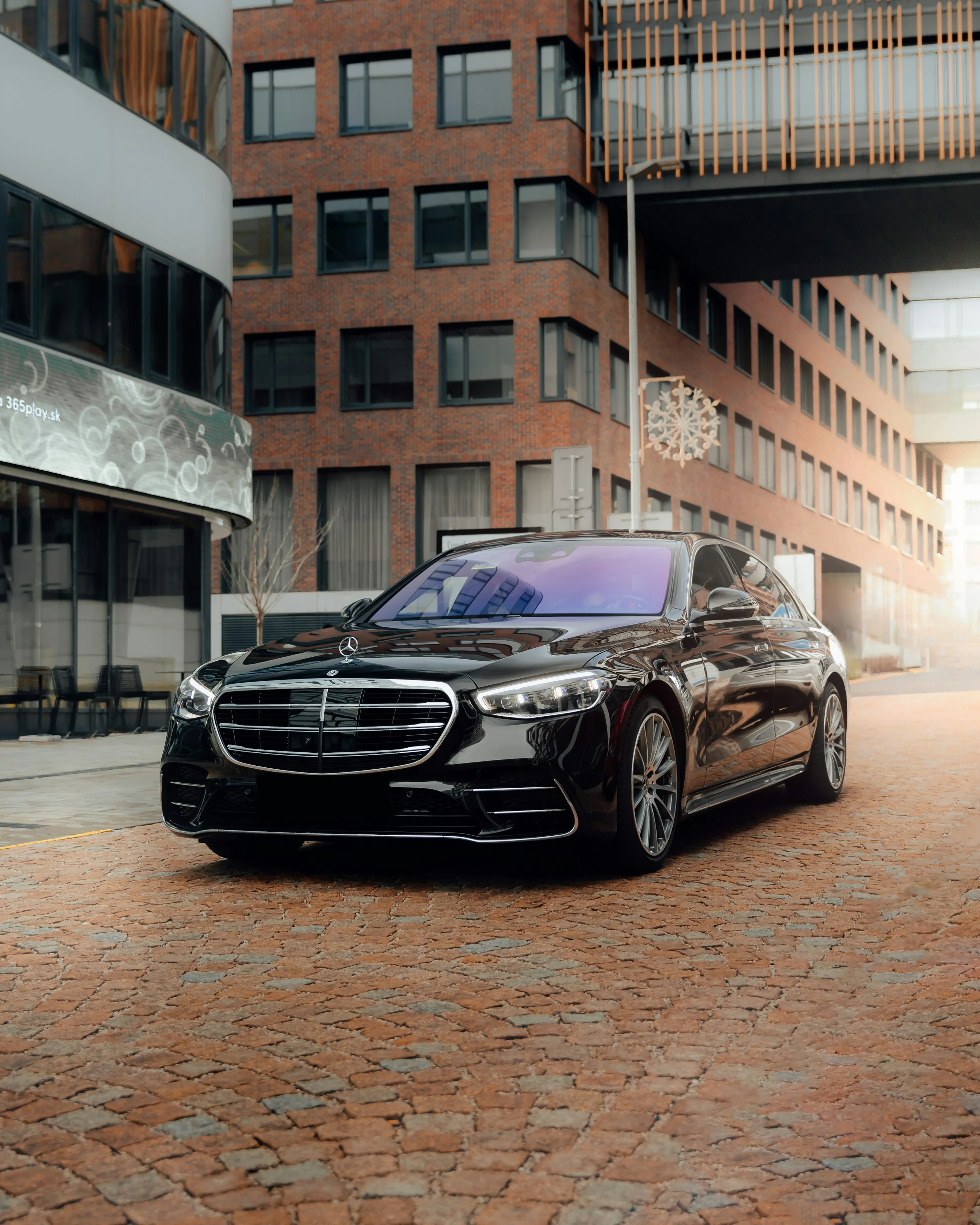 Mercedes-Benz Classe S noire rutilante de Kotewo, photographiée devant un immeuble moderne en briques rouges. Ce véhicule de luxe fait partie de notre flotte premium comprenant également les Mercedes Classe V, Classe E et Classe C, répondant à tous vos besoins de transport haut de gamme en région Rhône-Alpes. La berline arbore une finition noire étincelante, mettant en valeur sa calandre chromée distinctive et ses phares LED sophistiqués.