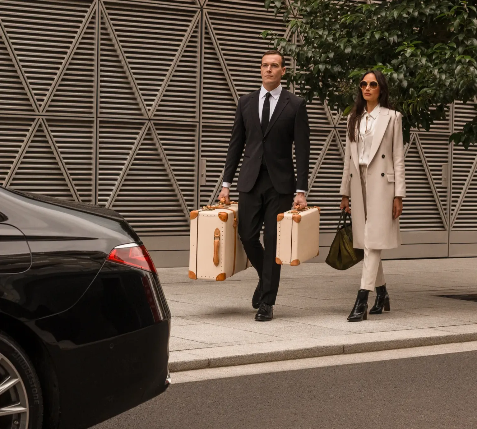 Service de chauffeur privé Kotewo en région Rhône-Alpes : Un chauffeur professionnel en costume noir porte des valises luxueuses en cuir clair, accompagnant une cliente élégante en ensemble blanc et long manteau beige vers une Mercedes berline noire. La scène se déroule devant un bâtiment moderne à l'architecture géométrique distinctive, symbolisant le service haut de gamme de Kotewo pour vos transferts aéroport et gare en Rhône-Alpes. Le professionnalisme du chauffeur et l'élégance du véhicule Mercedes illustrent l'engagement de Kotewo à offrir une expérience de transport premium et personnalisée.