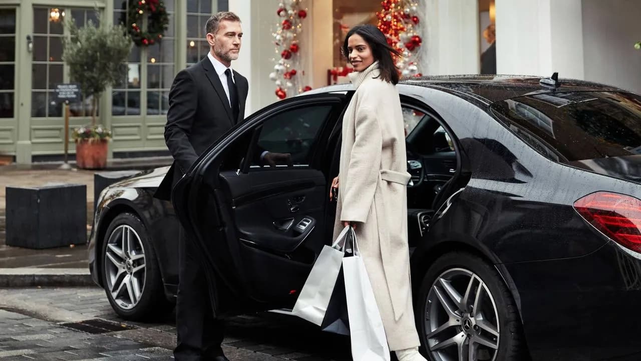 Service de chauffeur privé Kotewo en région Rhône-Alpes : Un chauffeur professionnel en costume noir ouvre la porte d'une Mercedes berline noire à une cliente élégante en manteau blanc, portant des sacs de shopping. La Mercedes, symbole de luxe et de confort, fait partie de la flotte haut de gamme proposée par Kotewo pour vos déplacements en Rhône-Alpes, que ce soit pour le shopping, les sorties ou les occasions spéciales.
