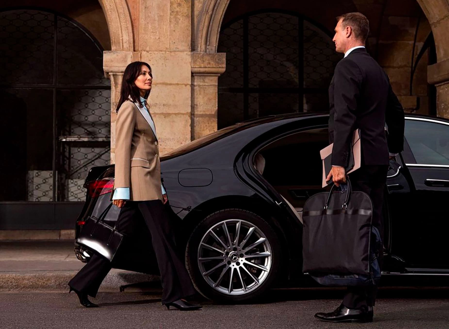 Service de chauffeur privé Kotewo en région Rhône-Alpes : Une femme d'affaires en tailleur beige et un homme en costume noir marchent vers une élégante Mercedes berline noire garée sous les arcades d'un bâtiment historique. Les deux personnes portent des bagages professionnels, illustrant un service de transport haut de gamme alliant confort et professionnalisme. La Mercedes, disponible en Classe C, E, S et V, symbolise l'excellence du service de chauffeur privé proposé par Kotewo dans toute la région Rhône-Alpes.